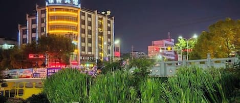 Un edificio de varios pisos con señalización iluminada, rodeado de vegetación y una calle con vehículos.