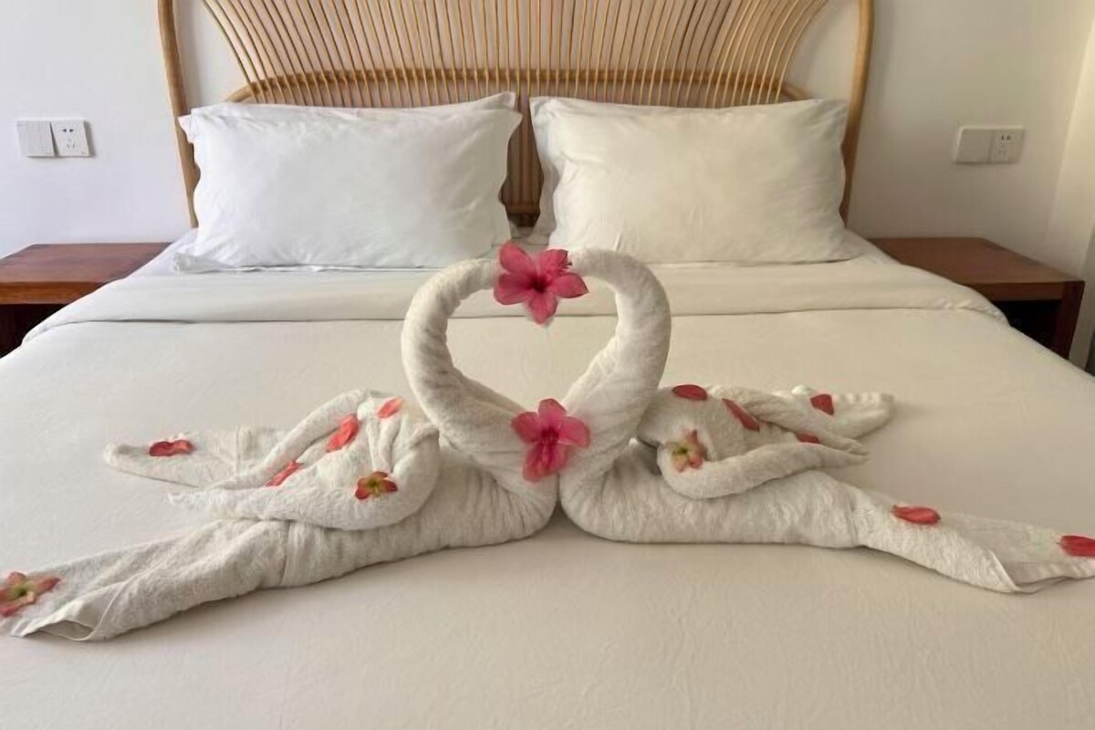 A bed with a white sheet, two pillows, and a towel artfully arranged in the shape of two swans with pink flowers.