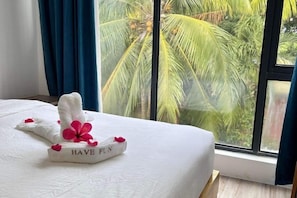 A neatly made bed with a towel and a flower arrangement on it, a window with a view of palm trees, and a blue curtain. - Fun Hotel (Panglao Municipality)