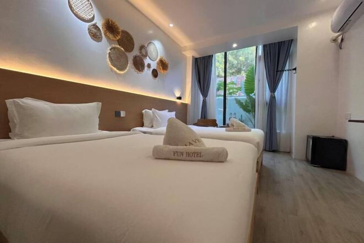A hotel room with two beds, a large window with curtains, a wall-mounted air conditioner, and a ceiling light fixture.