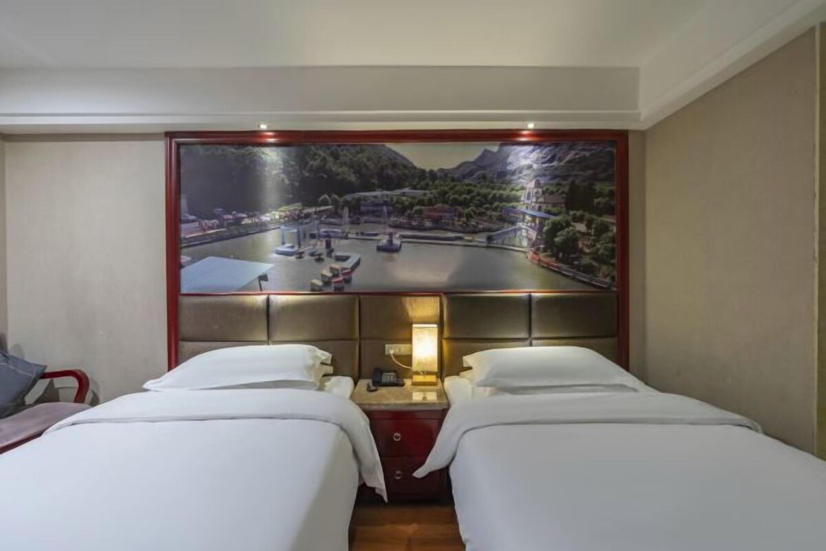 A hotel room with two beds, a large scenic mural on the wall, a bedside table with a lamp, and a small desk with a chair.