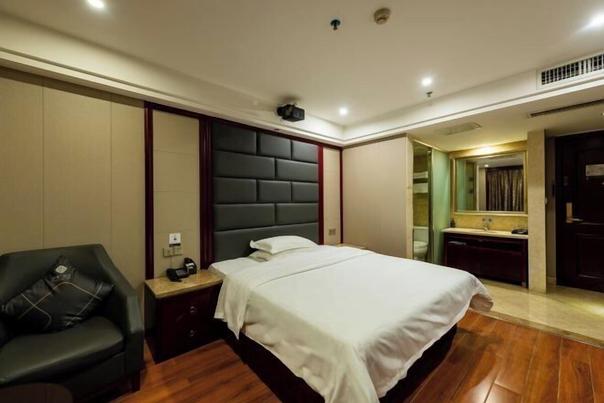 A hotel room with a large bed, a dark leather sofa, a nightstand with a phone, and a bathroom visible through an open door.