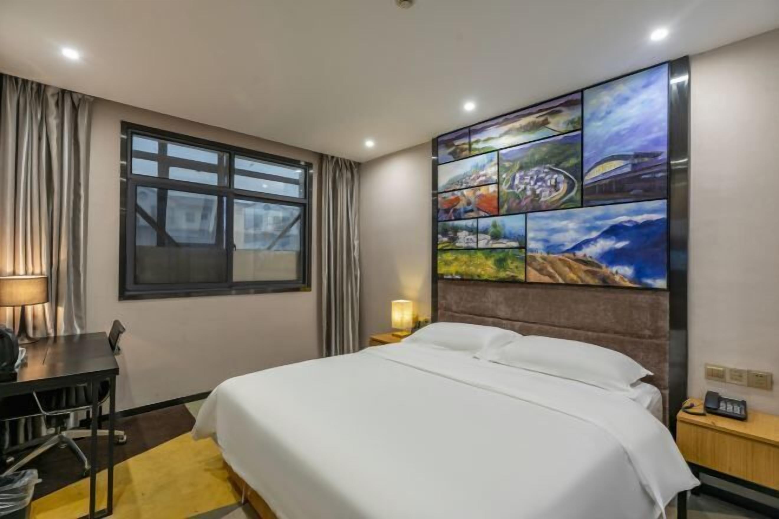 Habitación de hotel con cama, escritorio, silla, lámpara, mesita de noche y un cuadro de un paisaje urbano en la pared.