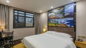 Habitación de hotel con cama, escritorio, silla, lámpara, mesita de noche y un cuadro de un paisaje urbano en la pared.