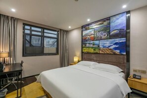 A hotel room with a bed, a desk, a chair, a lamp, a nightstand, and a wall with a cityscape painting.