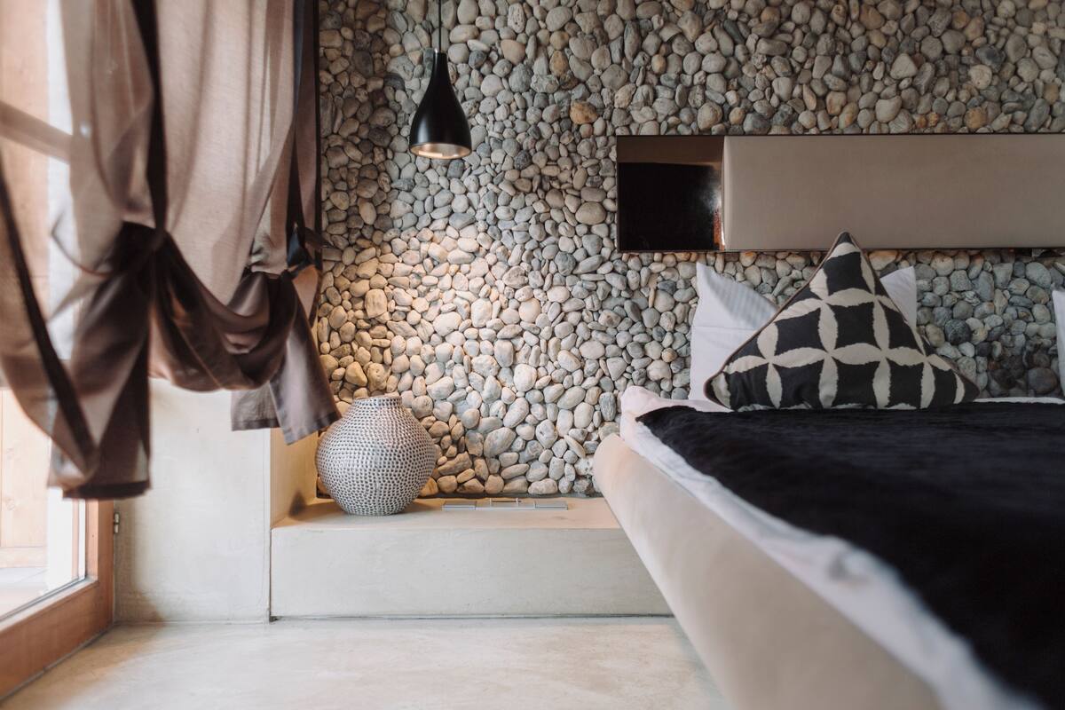 A bedroom with a bed, a headboard, a wall of pebbles, a hanging light, and a window with curtains.