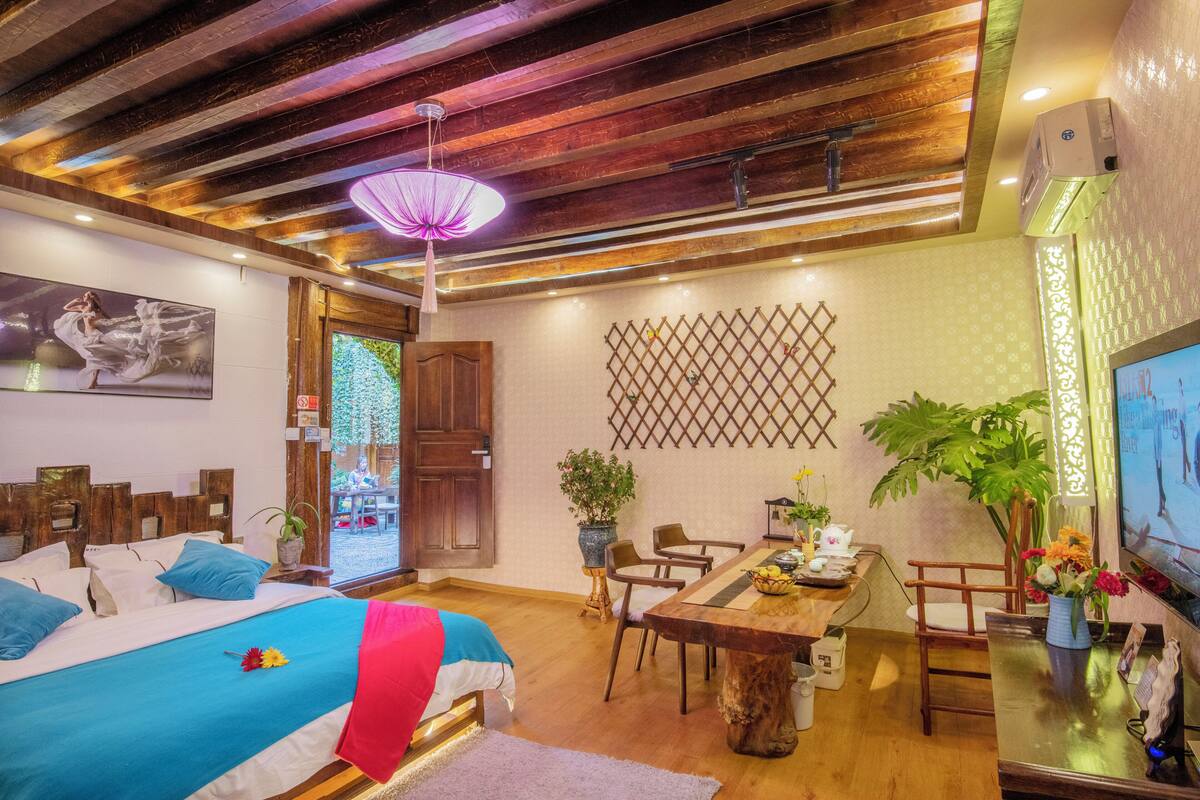 A well-lit room with a bed, wooden ceiling, a dining table with chairs, a TV, and a potted plant.