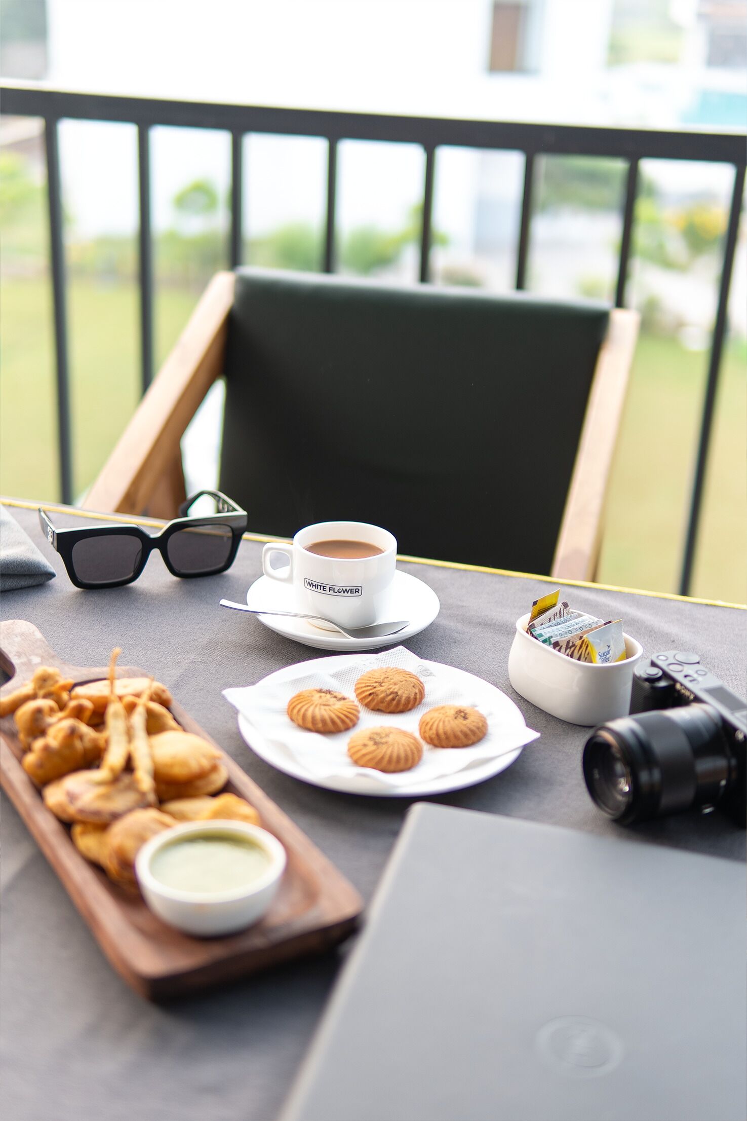 A table with a cup of coffee, sunglasses, pastries, a camera, and a laptop on a balcony.