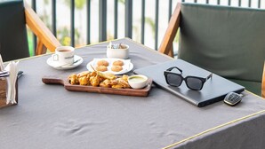 Une table est dressée sur un balcon, d’où l’on aperçoit un immeuble et de la verdure. On y trouve un ordinateur portable, des lunettes, un téléphone portable, une assiette de biscuits, une tasse de café et un bol de soupe.