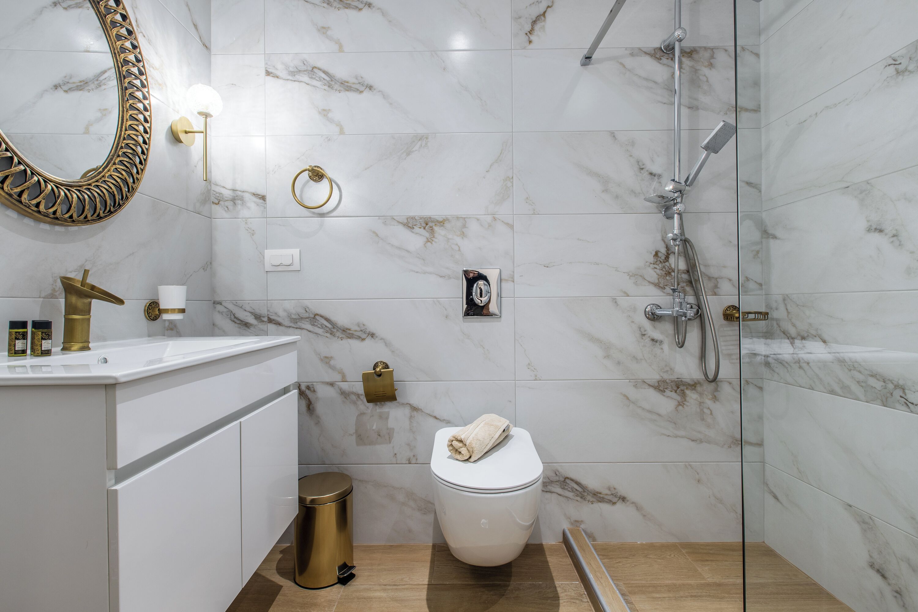 A modern bathroom with a marble floor, a white toilet, a gold trash bin, a glass shower enclosure, and a large mirror with a gold frame.