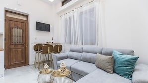 A modern living room with a grey sofa, a wooden door, a dining table with chairs, and a TV mounted on the wall.