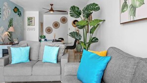 Una sala de estar con un sofá gris, cojines azules y amarillos, un ventilador de techo y plantas verdes grandes.