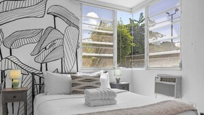 Un dormitorio con cama, mesita de noche, lámpara, radiador y ventanas amplias que dan a árboles y una valla.