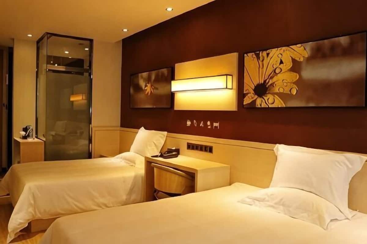 A hotel room with two beds, a bedside table with a lamp, a mirror, and artwork on the wall.