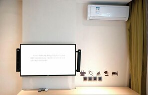 A hotel room with a mounted TV, air conditioning unit, and a wall with various icons indicating amenities like Wi-Fi, power outlets, and a coffee machine.
