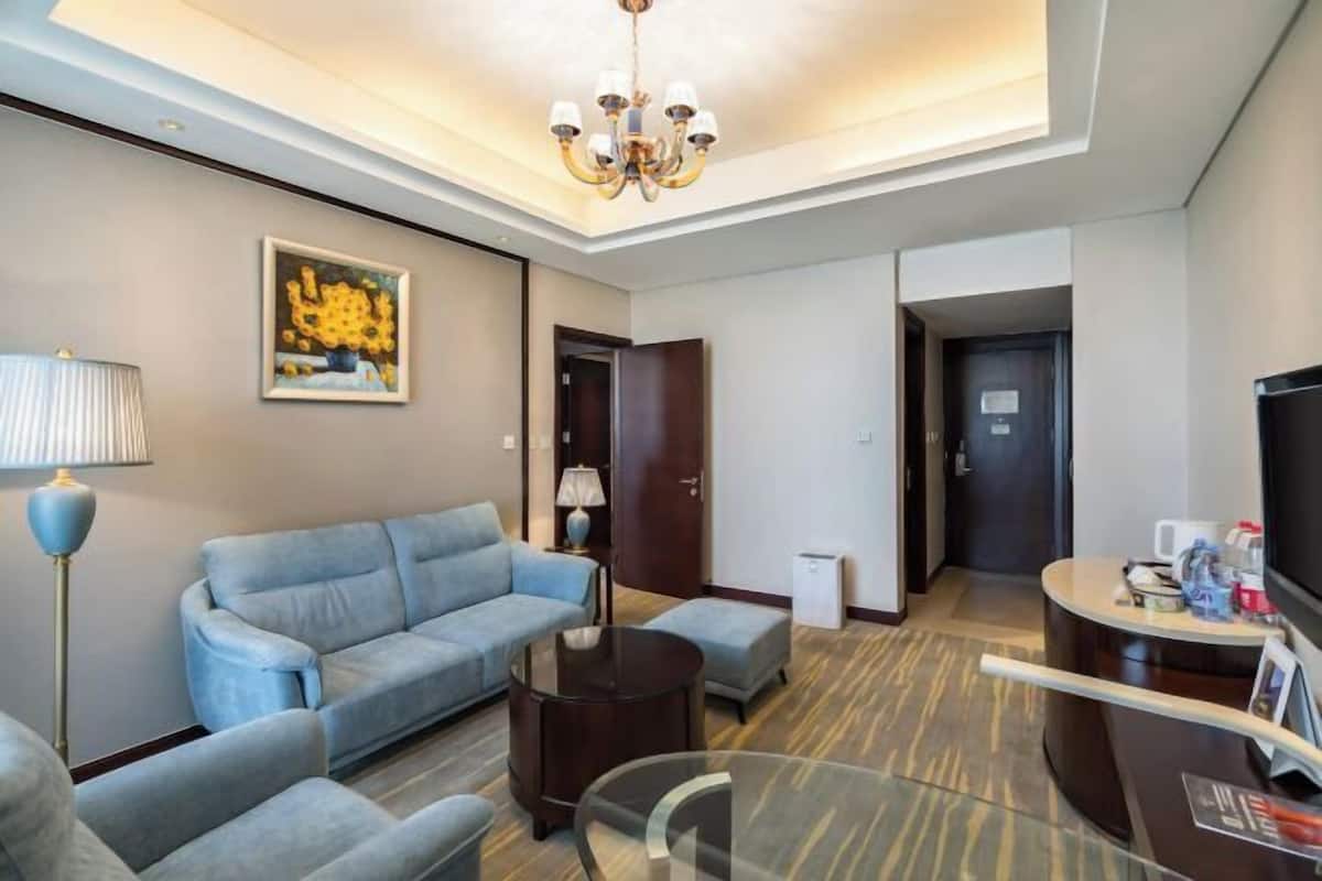 A modern hotel room with a sofa, armchairs, a glass coffee table, a desk with a computer, and a television.