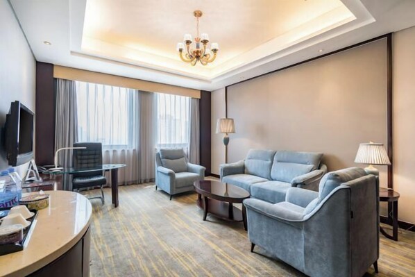 A hotel room with a sofa, armchairs, a TV, a desk, and a chandelier.