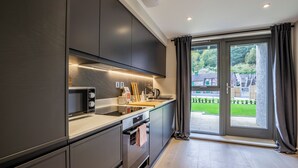 Uma cozinha moderna com armários escuros, micro-ondas, cafeteira e vista para um jardim verdejante através da porta de vidro.