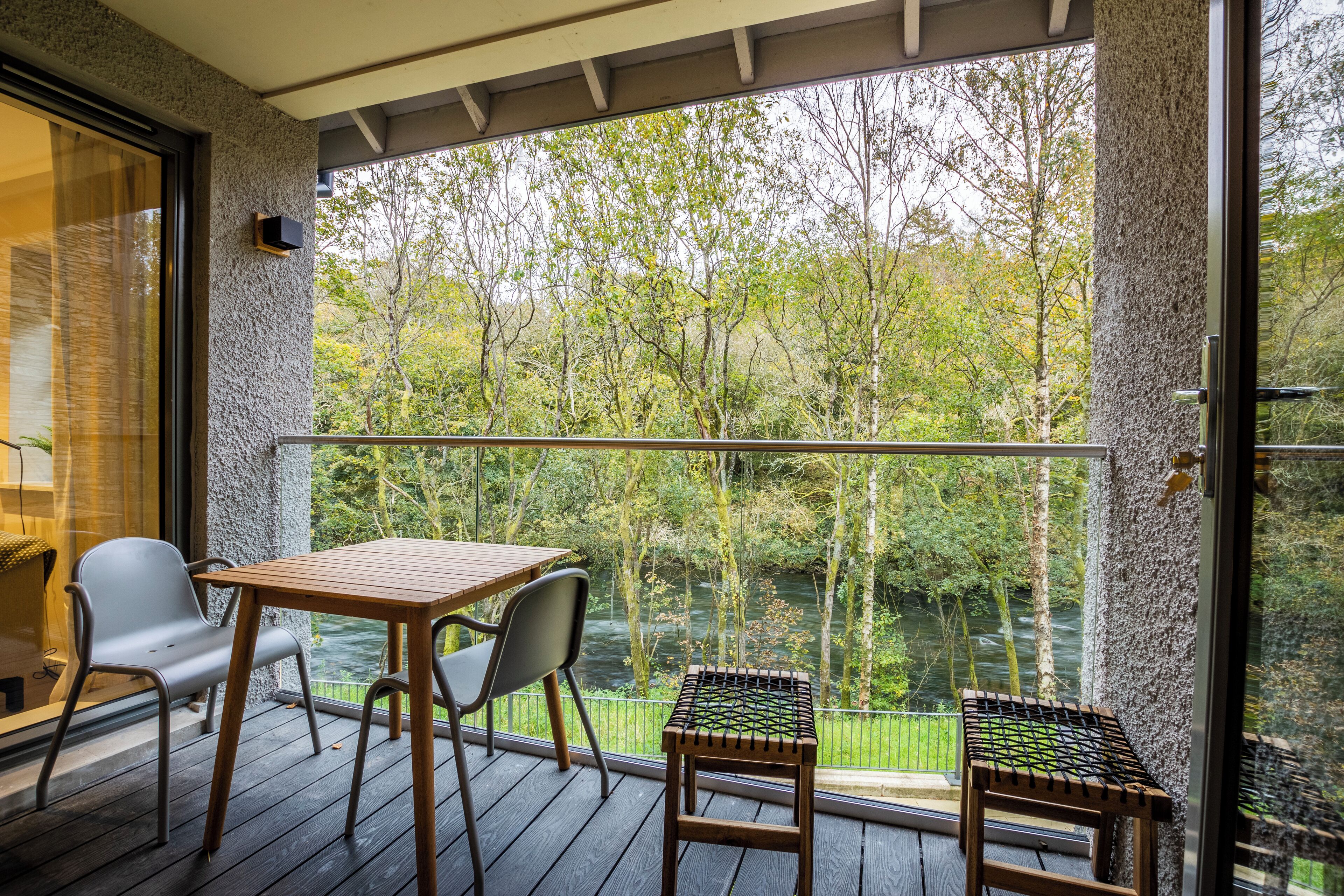Un balcón con mesa y sillas de madera, un taburete pequeño y vistas a un río y árboles.