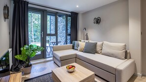Uma sala de estar moderna com um sofá, uma mesa de centro com frutas, uma planta em vaso e vista do exterior através de grandes janelas.