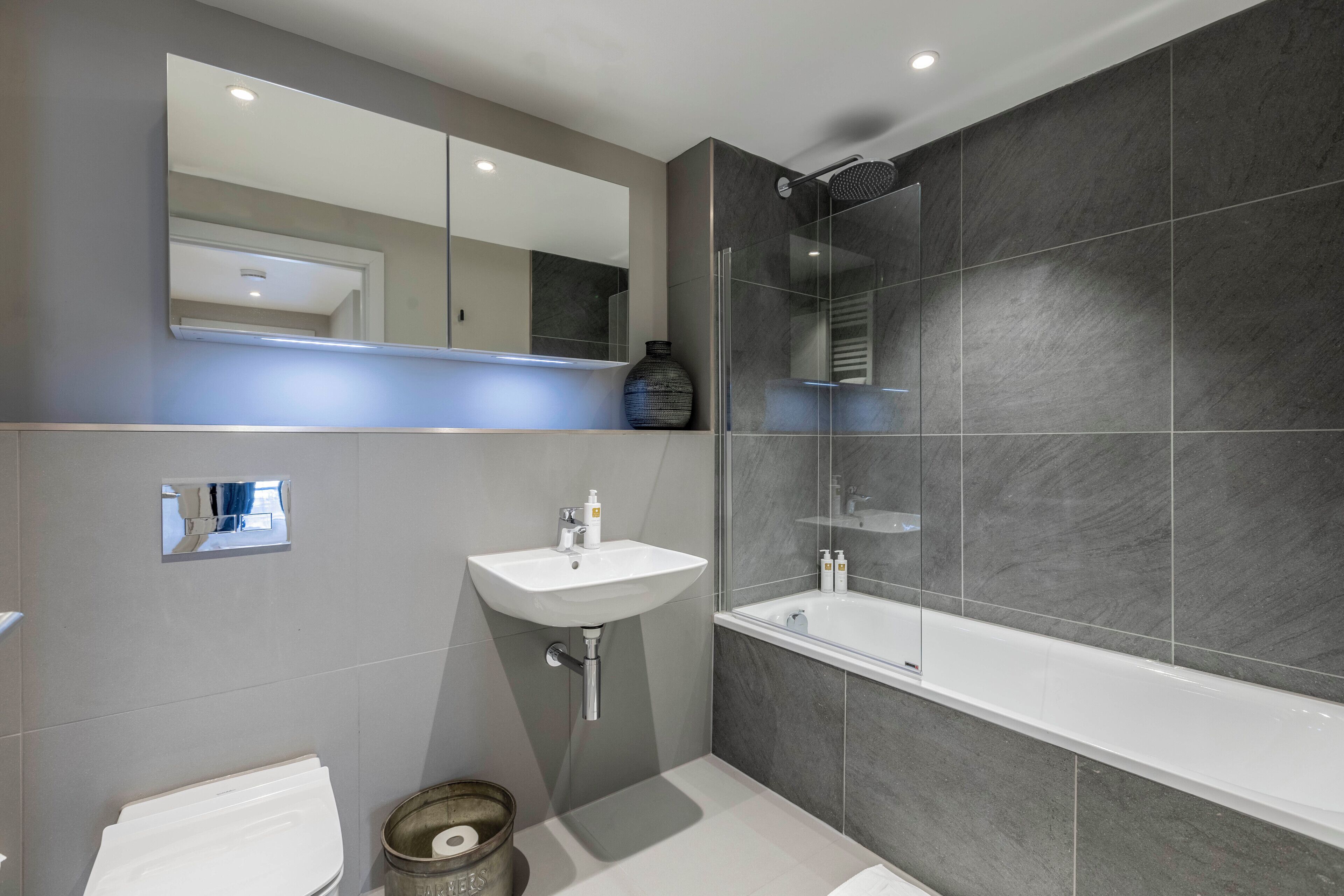 A modern bathroom with a large mirror, a white sink, a bathtub, and dark grey tiles.
