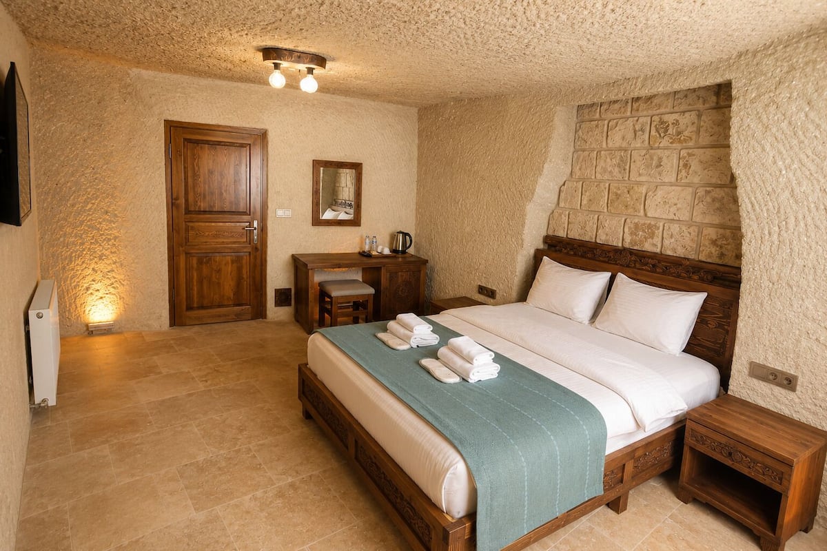 A bedroom with a wooden bed, a nightstand, a desk, a chair, and a stone wall.
