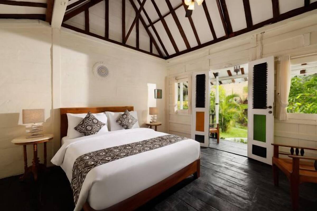 Een slaapkamer met een groot bed, een houten hoofdbord, nachtkastjes en uitzicht op een tuin door een open deur.