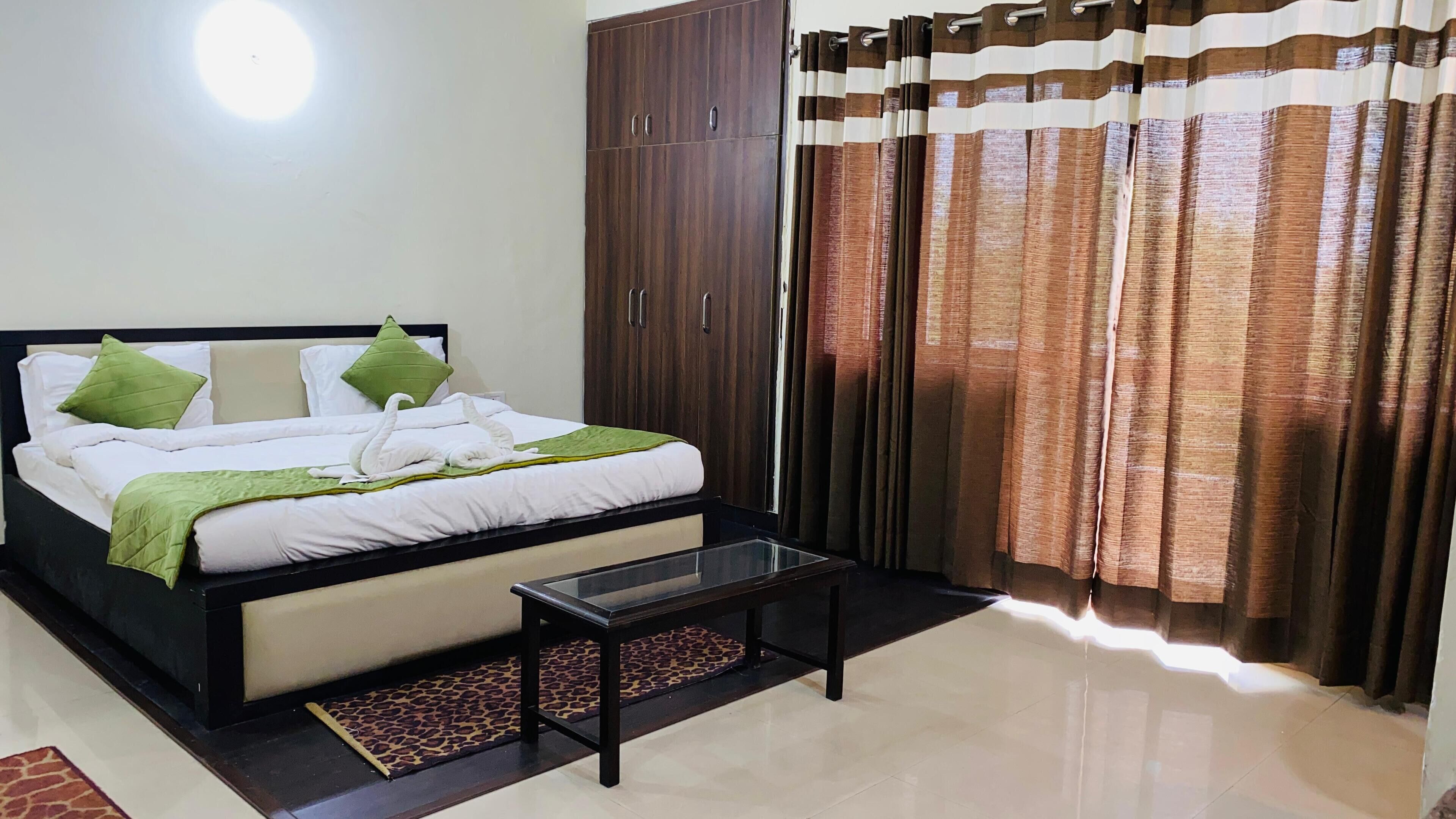 Una habitación de hotel con cama, almohadas verdes, una mesa de centro de vidrio, un armario y cortinas.