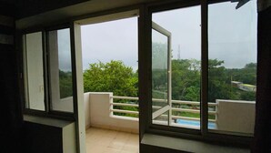 A room with a large window overlooking a balcony with a railing and a view of greenery and a distant tower. - LivBox Rudrapur (Kichha)