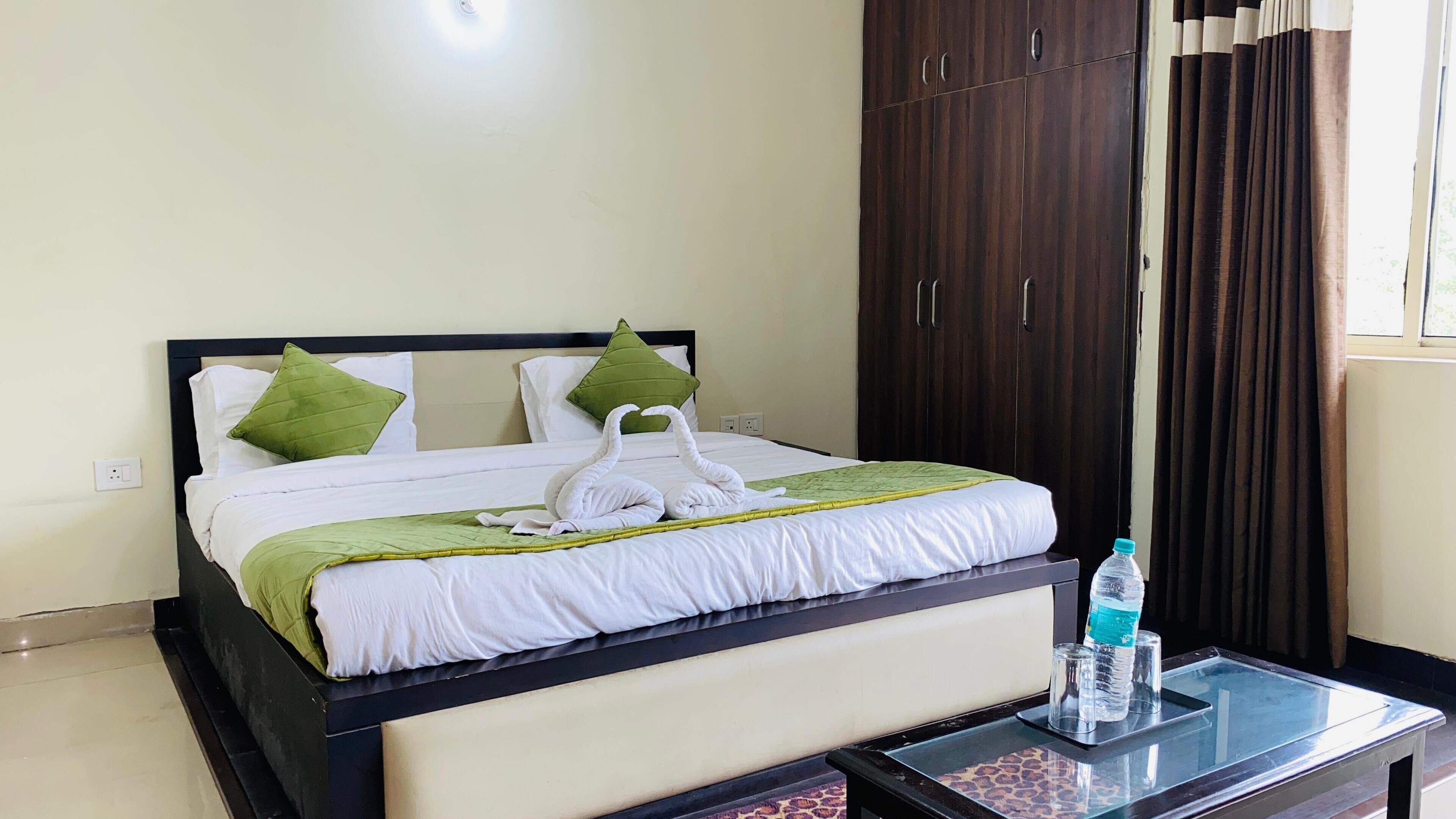Una cama bien hecha con ropa de cama blanca y verde, una mesita de vidrio con botellas de agua y un armario de madera oscura.
