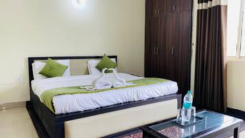 Una cama bien hecha con ropa de cama blanca y verde, una mesita de vidrio con botellas de agua y un armario de madera oscura.