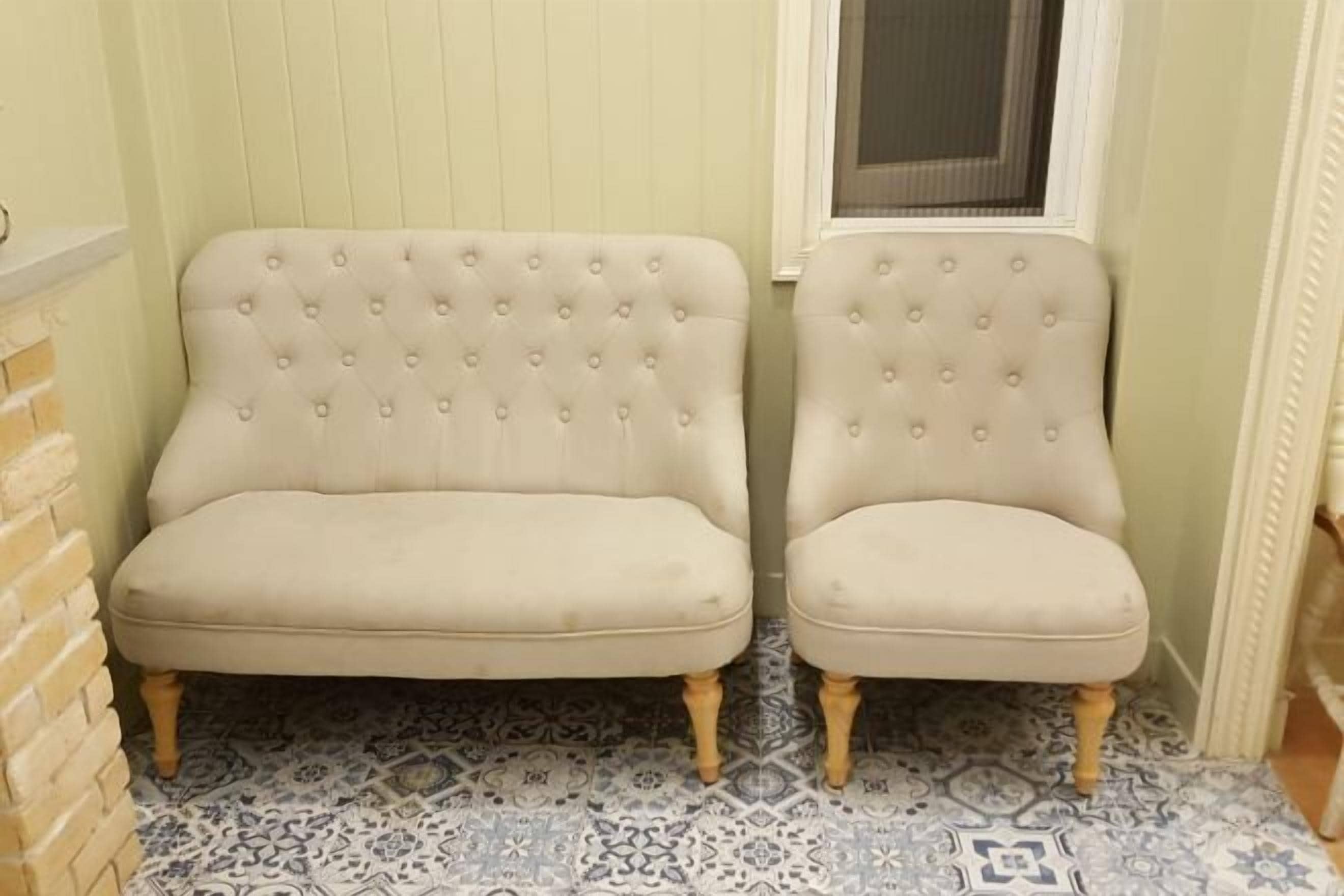 Duas cadeiras estofadas bege com pernas de madeira, posicionadas sobre ladrilhos estampados num canto com janela e porta.