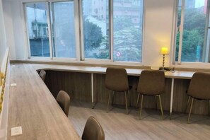 A room with a long wooden desk, four chairs, and a window with a view of buildings and trees. - TW Hostel 2 (Taipei)