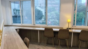 Una habitación moderna con un escritorio de madera largo, cuatro sillas marrones y un ventanal con vistas a edificios y árboles.