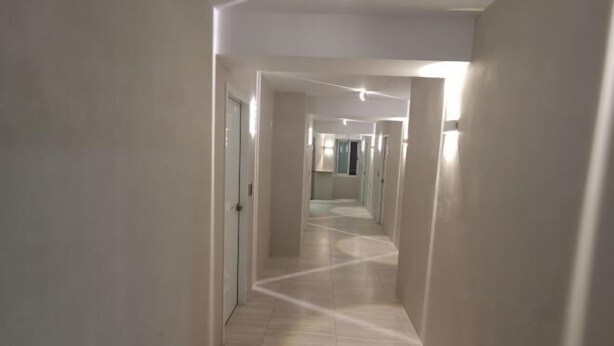 Un pasillo largo y estrecho con paredes blancas y suelo de tonos claros, que conduce a una habitación con ventana y puerta.
