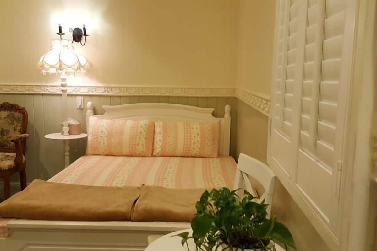 Una cama con una colcha estampada y almohadas, una mesita de noche con una lámpara, una silla y una planta en maceta.