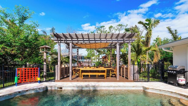 Une piscine aux eaux cristallines, surmontée d’une pergola en bois, sous laquelle se trouvent une table et des chaises.