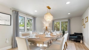 Un coin repas avec une table en bois dressée pour six personnes, entourée de chaises blanches, d’un lustre et de grandes fenêtres ornées de rideaux.