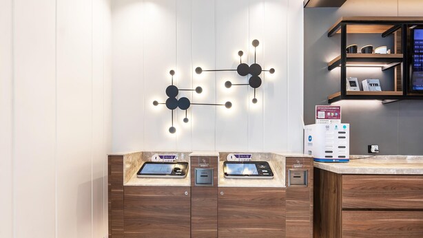 A modern hotel reception desk with digital check-in kiosks, a wall-mounted light fixture, and a small shelf with items. - Hanting Hotel (Zhengzhou)