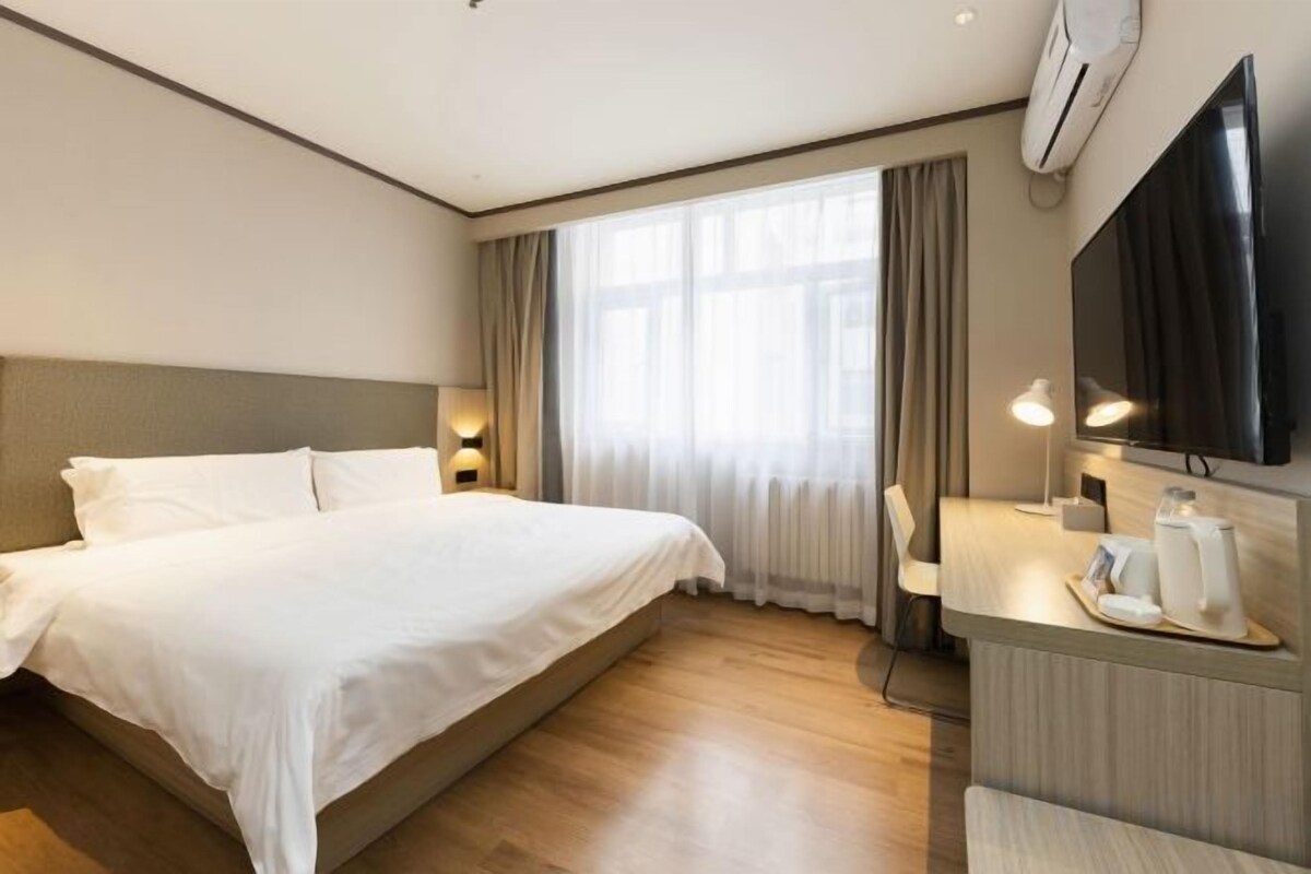 A hotel room with a large bed, a desk with a chair, a flat-screen TV, and a window with curtains.