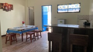 A dining area with a table set for four, wooden stools, a small refrigerator, and a window with a floral curtain. - Carolina Townhome (Catanauan)