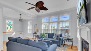 A spacious living room with a large grey sofa, a coffee table, and a fireplace. There are large windows allowing natural light and a ceiling fan above. - Palmetto Moon | Ocean Point Retreat near Beach, Pool & Golf (Isle of Palms)