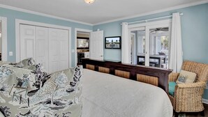 A bedroom with a large bed, a wicker chair, a TV, and a view of the outside. - Palmetto Moon | Ocean Point Retreat near Beach, Pool & Golf (Isle of Palms)