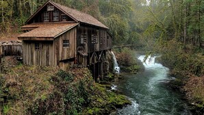 A rustic wooden house with a steeply pitched roof, situated on stilts over a flowing river, surrounded by dense forest. - Barnehagen (Los Alamos)