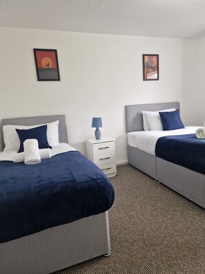Une chambre d’hôtel avec deux lits, chacun doté d’une tête de lit grise, de draps blancs et d’une couverture bleue. On y trouve une table de chevet blanche avec une lampe, et un tableau encadré est accroché au mur.