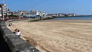 A sandy beach with a curved stone wall, a few people sitting on the wall, and buildings in the background. - Seasideescape-2br-sleeps4-parking-petsok-fireplace (Bridlington)