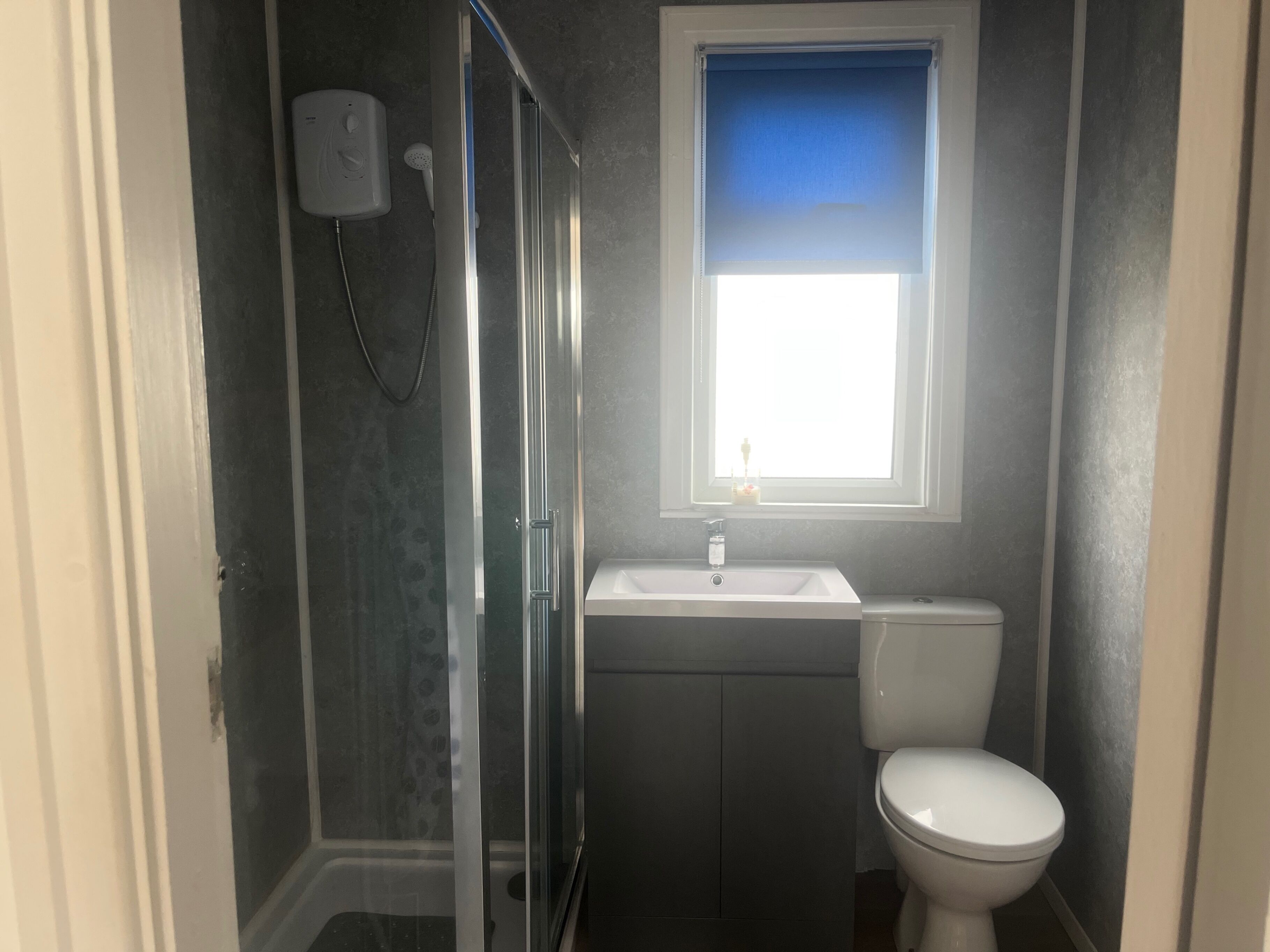 A bathroom with a glass shower enclosure, a white toilet, and a window with a blue blind.