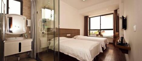 A hotel room with a glass shower enclosure, a bed with a wooden headboard, a bedside table, and a window with a view.