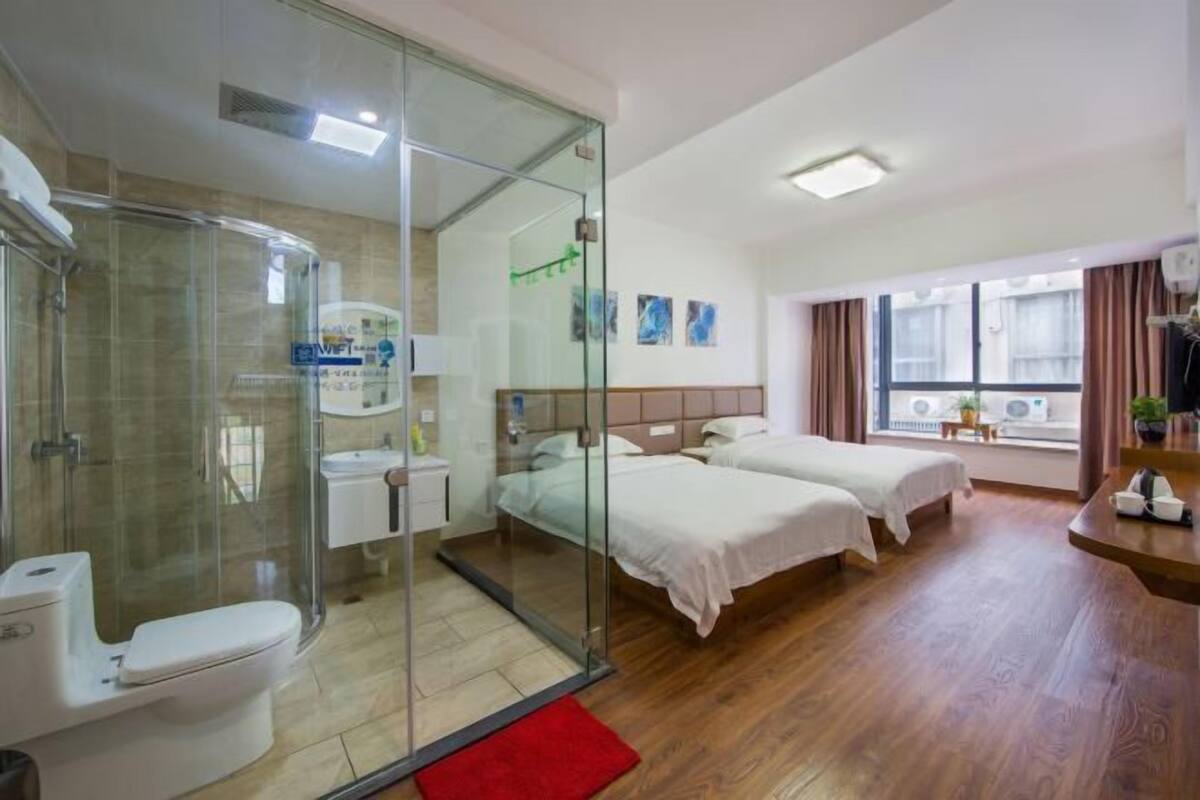 A hotel room with a glass shower enclosure, a bed with two pillows, a bedside table, and a window with curtains.