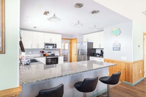 A modern kitchen with granite countertops, stainless steel appliances, and a central island with bar stools. - Third Row in Whalehead with Pool + Spa, Sleeps 22! (Corolla)